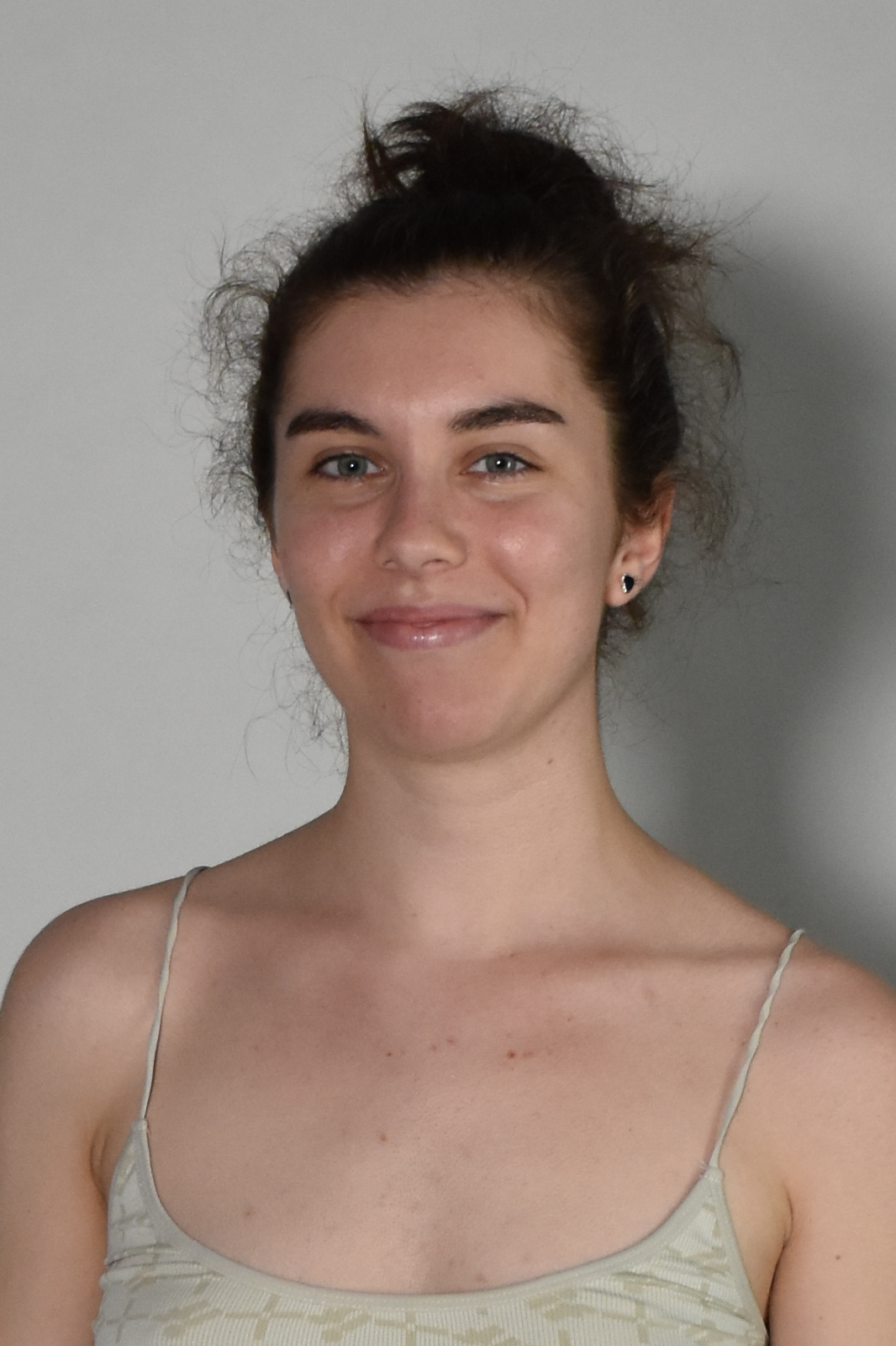Smiling young woman with dark hair in a messy bun, wearing a light patterned tank top against a neutral background.
