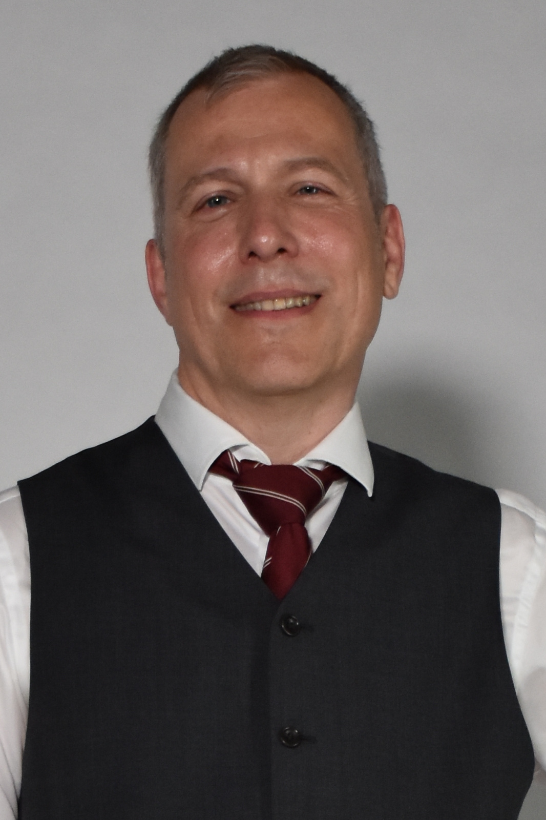 Smiling man in a white shirt, charcoal vest, and red striped tie against a plain light background (professional head-and-shoulders portrait).