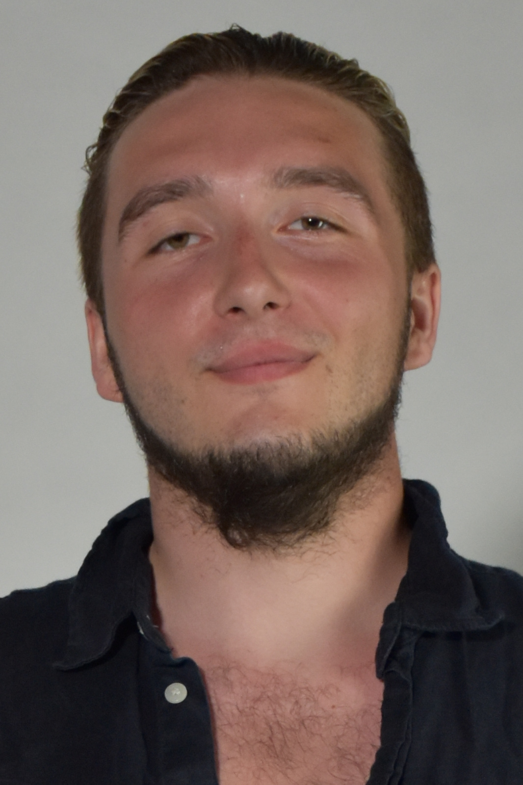 Close-up portrait of a young man with short dark hair and light beard, wearing a dark button-up shirt, looking at the camera with a slight smile against a plain background.