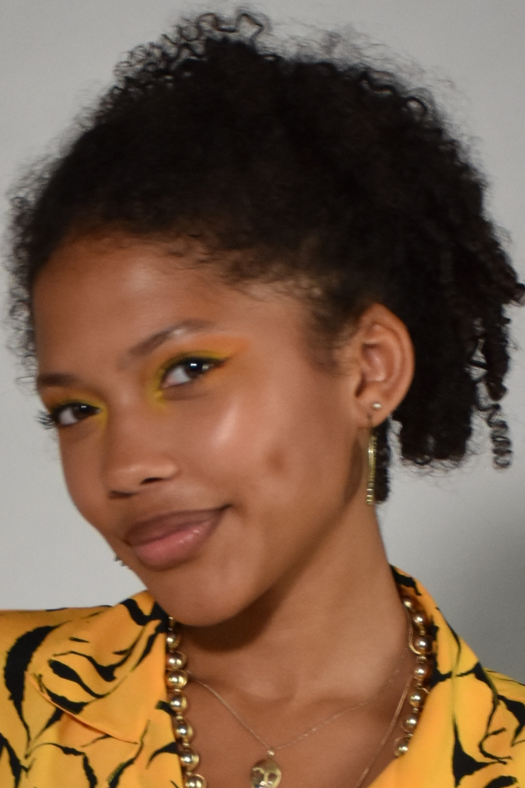 Young woman with short curly hair wearing a yellow patterned top, yellow eye makeup, gold jewelry, and a gentle smile.