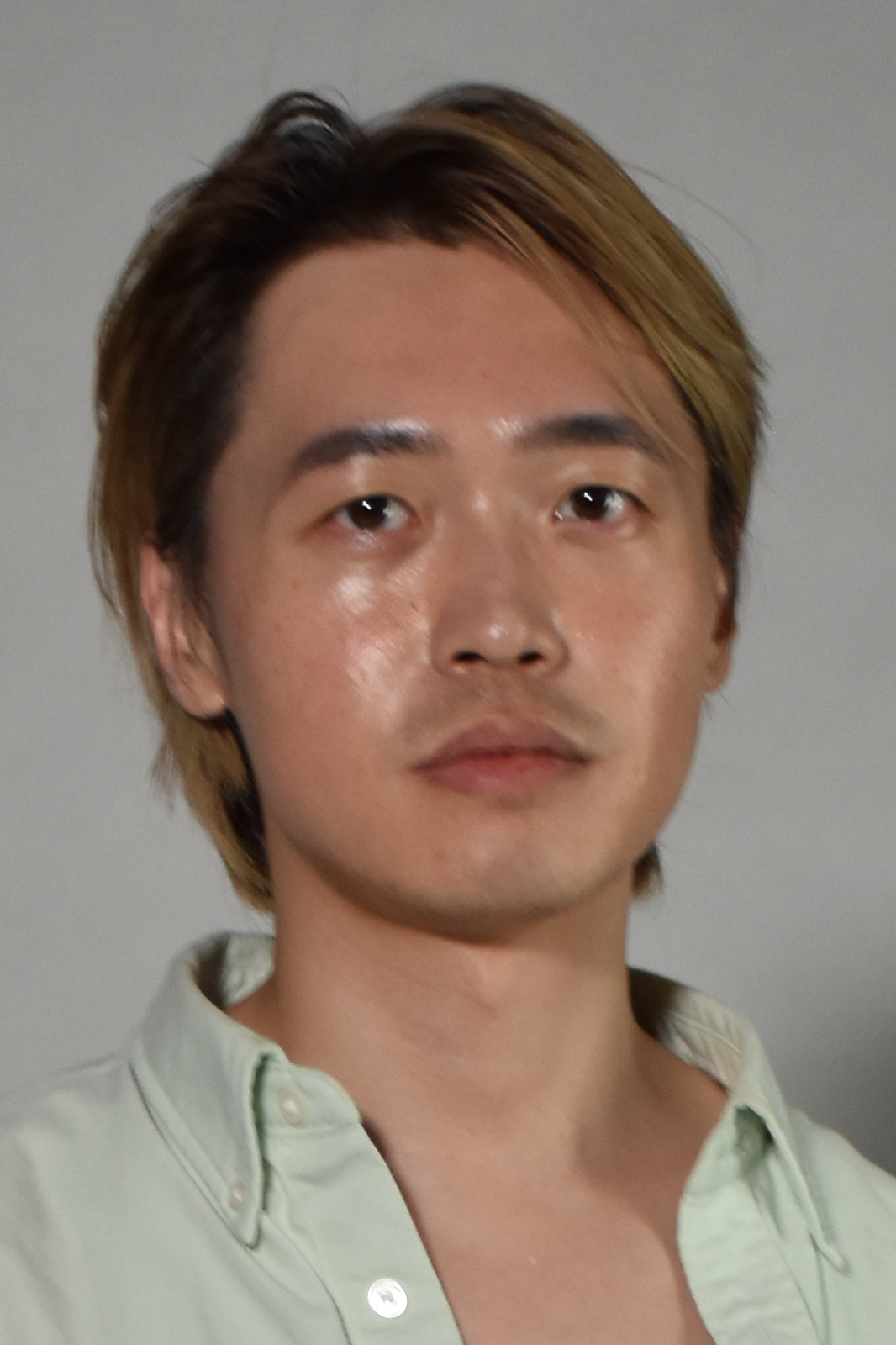 Portrait of an Asian man with blonde hair in a pale green shirt, neutral expression, gray studio background.