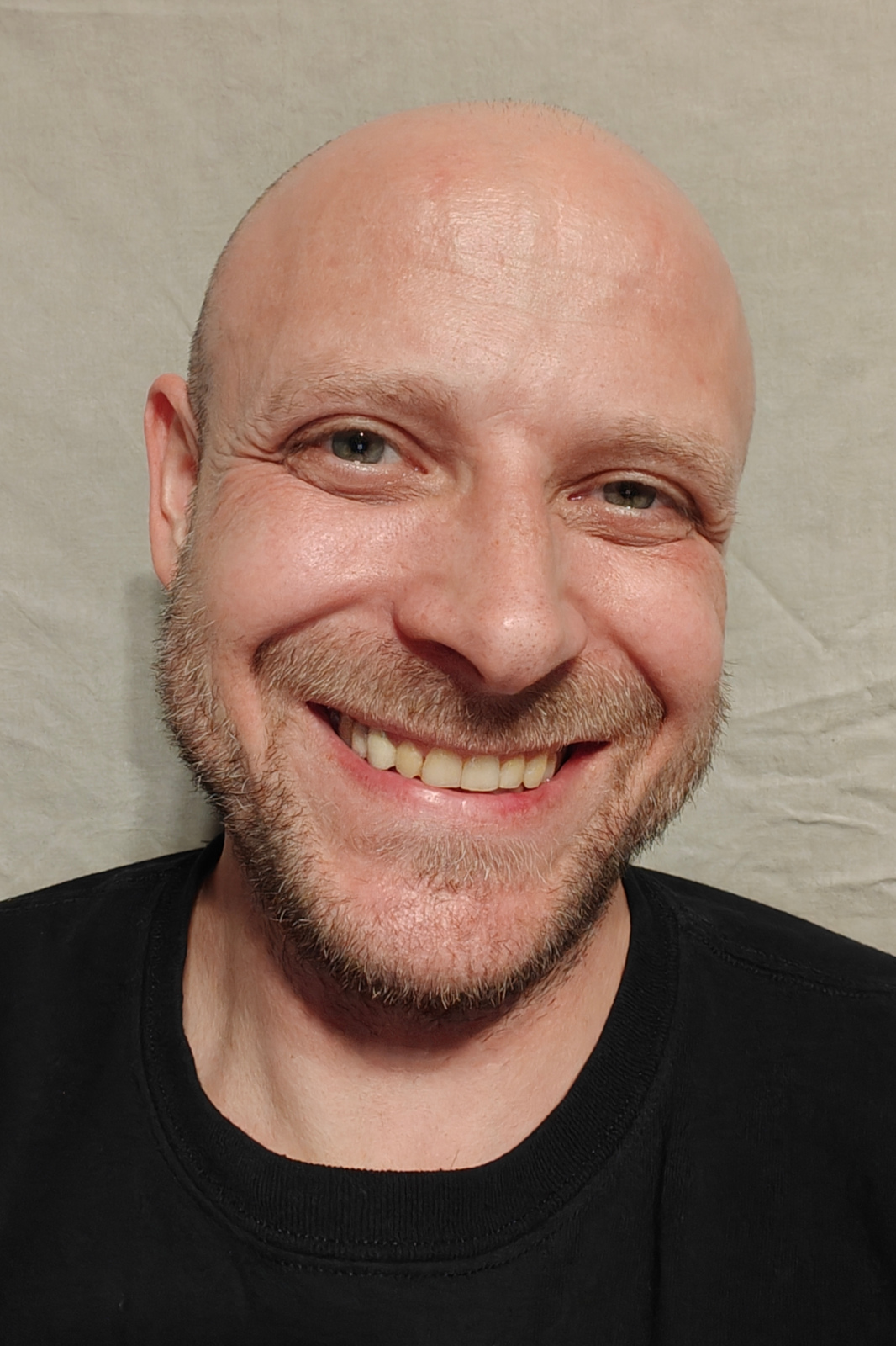 JohannesWebsitePortrait - Rainbow Gold Kulturverein - Rainbow Gold Kulturverein Close-up portrait of a smiling bald man with light stubble, wearing a black shirt, neutral backdrop behind him.