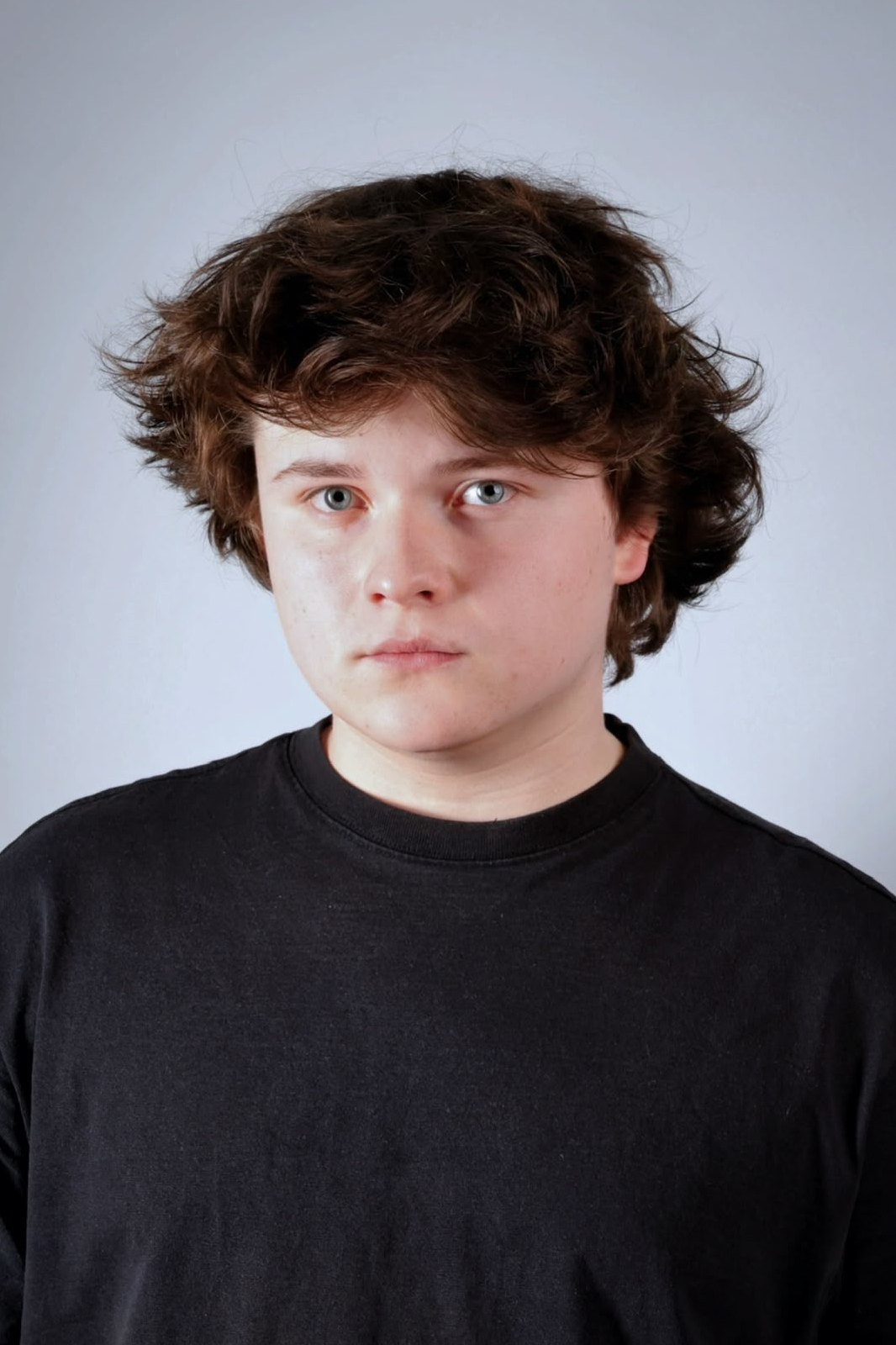 SimonWebsitePortrait - Rainbow Gold Kulturverein - Rainbow Gold Kulturverein Close-up portrait of a young person with tousled brown hair, wearing a black shirt, looking straight at the camera against a pale gray background.